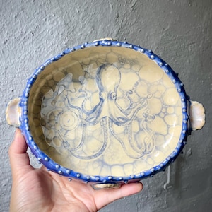 Op de afbeelding: Een ovale keramische kom met een crèmekleurige binnenkant voorzien van een blauwe octopusillustratie en een bellenpatroon. De buitenrand is getextureerd, donkerblauw met verhoogde stippen, en bevat twee handvatten en kleine schelpvormige accenten.