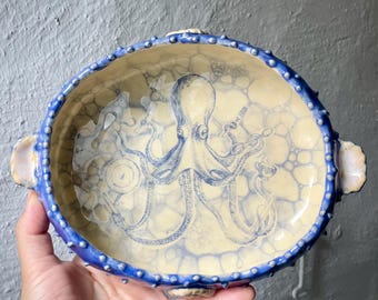 Puede incluir: Un cuenco ovalado de cerámica con interior color crema que presenta una ilustración de pulpo azul y un patrón de burbujas. El borde exterior es texturizado, azul oscuro con puntos en relieve, e incluye dos asas y pequeños detalles de conchas.