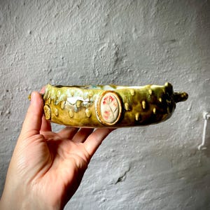 Puede incluir: Un cuenco de cerámica redondo con un esmalte verde oliva texturizado y abultado, con dos pequeñas asas. El exterior presenta un medallón ovalado central que muestra una delicada ilustración floral rosa. El borde tiene una forma orgánica irregular.