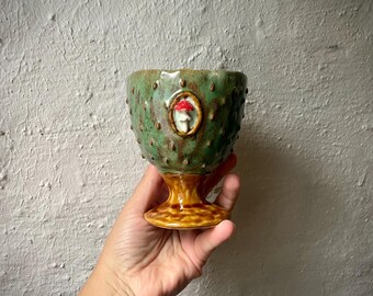 Puede incluir: Una copa de cerámica con un cuenco verde texturizado con protuberancias en relieve y un pequeño medallón ovalado central que muestra un hongo blanco con sombrero rojo. La copa tiene una base de pedestal marrón lisa y brillante con textura.