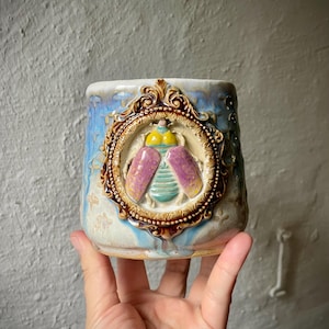 Könnte beinhalten: Eine Keramik-Tasse mit einem detaillierten Relief einer Insektenfigur in einem verzierten, goldfarbenen Rahmen. Die Tasse hat einen blau-cremefarbenen Hintergrund mit strukturiertem Finish. Der Käfer hat lila Flügel und einen gelben Kopf.