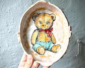Puede incluir: Un cuenco de cerámica redondo con borde festoneado que presenta una ilustración pintada a mano de un oso de peluche marrón con pantalones cortos de color verde azulado y una pajarita roja. El interior es crema con un borde marrón.