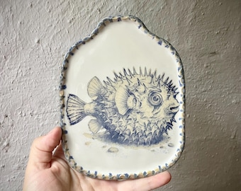 Puede incluir: Una bandeja de cerámica con esmalte blanco roto y una ilustración azul dibujada a mano de un pez globo en el centro. La bandeja tiene una forma rectangular redondeada irregular con un borde festoneado decorativo en azul y beige.