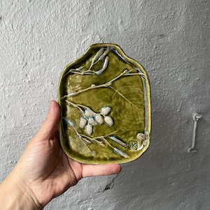 Könnte beinhalten: Eine handgefertigte, olivgrüne Keramikschale mit rechteckiger Form und erhöhtem Rand. Die Schale zeigt ein detailliertes botanisches Design aus Zweigen und Beeren in einer helleren grünen und weißen Glasur. Die Schale ist etwa 15 cm lang.