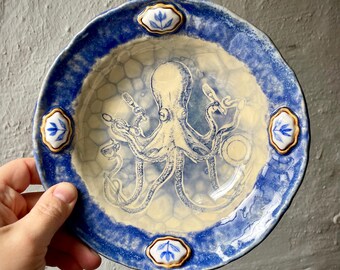 Puede incluir: Un cuenco de cerámica redondo con un centro de color crema que presenta una ilustración de un pulpo azul oscuro sosteniendo varios utensilios. El borde es azul texturizado con cuatro detalles blancos en forma de diamante con motivos florales.