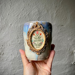 Può includere: Una tazza in ceramica con finitura smaltata marmorizzata blu e bianca. La parte anteriore presenta una cornice ovale dorata in rilievo contenente un'illustrazione botanica dipinta di uno stelo centrale con fiori rossi e piccoli fiori blu e rosa.