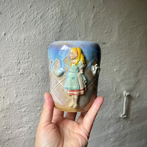 Könnte beinhalten: Eine Keramiktasse mit einem Relief eines Mädchens in blauem Kleid und Schürze. Die Tasse hat ein Rautenmuster und eine blaue und beige Glasur. Die Tasse wird in einer Hand gehalten.