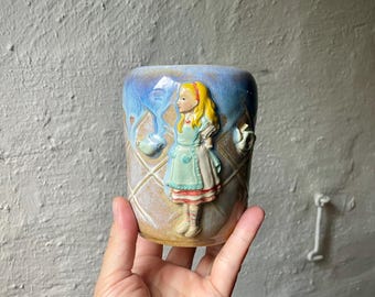 Puede incluir: Una taza de cerámica con un relieve de una niña con vestido azul y delantal. La taza tiene un patrón de diamantes y un esmalte azul y beige. La taza está sujeta con una mano.