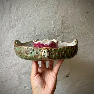 Könnte beinhalten: Eine Keramikschale mit strukturierter grüner Außenseite, die mit erhabenen Noppen und einem zentralen ovalen Blumenemblem versehen ist. Das Innere ist hell mit kleinen Sprenkeln und enthält eine kleinere, gerüschte, magentafarbene Schale.