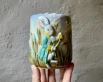 Puede incluir: Una taza de cerámica con un relieve tridimensional de un conejo blanco con abrigo azul y chaleco amarillo, sosteniendo un reloj de bolsillo dorado. El fondo muestra hierba alta verde y un cielo azul claro, con un acabado brillante.