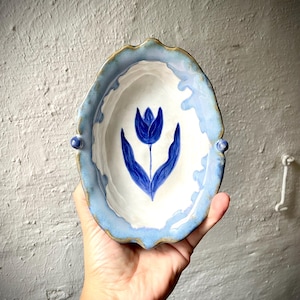 Könnte beinhalten: Eine ovale Keramikschale mit gewelltem Rand und zwei kleinen Seitengriffen. Das Innere ist weiß und mit einem handgemalten, dunkelblauen Tulpenmotiv versehen. Der Rand ist hellblau glasiert und mit braunen Akzenten verziert.