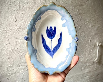 Puede incluir: Un cuenco ovalado de cerámica con borde festoneado y dos pequeñas asas laterales. El interior es blanco con un diseño de tulipán azul oscuro pintado a mano. El borde presenta un esmalte azul claro con detalles marrones decorativos.