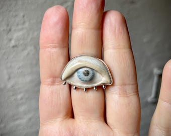 Puede incluir: Un anillo único con un diseño de ojo. El anillo tiene una banda de color plateado y un ojo detallado con un iris azul claro y una pupila oscura. El ojo está sobre un fondo color crema. El anillo tiene pequeños detalles plateados debajo.