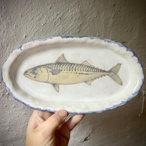 Könnte beinhalten: Eine ovale Servierplatte aus Keramik mit weiß glasierter Oberfläche und einem handbemalten, blauen, gewellten Rand. In der Mitte der Platte befindet sich eine detaillierte Illustration einer Makrele in Gelb-, Grau- und Schwarztönen.