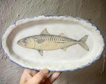 Puede incluir: Una bandeja de servicio ovalada de cerámica con una superficie esmaltada en blanco y un borde festoneado pintado a mano en azul. El centro de la bandeja presenta una ilustración detallada de una caballa en tonos amarillos, grises y negros.
