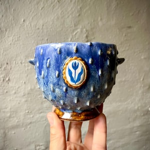 Puede incluir: Un cuenco de cerámica azul con una superficie texturizada de púas y una pequeña asa a cada lado. El cuenco presenta un medallón ovalado central con una ilustración de un tulipán azul sobre fondo blanco, enmarcado por un borde dorado festoneado, sobre una base marrón.