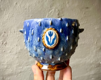 Puede incluir: Un cuenco de cerámica azul con una superficie texturizada de púas y una pequeña asa a cada lado. El cuenco presenta un medallón ovalado central con una ilustración de un tulipán azul sobre fondo blanco, enmarcado por un borde dorado festoneado, sobre una base marrón.