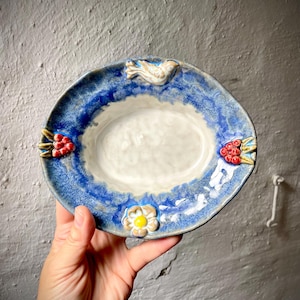 Könnte beinhalten: Eine ovale Keramikschale mit cremefarbener Mitte und strukturiertem blauem Rand. Der Rand ist mit einem kleinen modellierten Vogel oben, einer einzelnen weißen Blume mit gelber Mitte unten und roten Beerenclustern an den Seiten verziert.