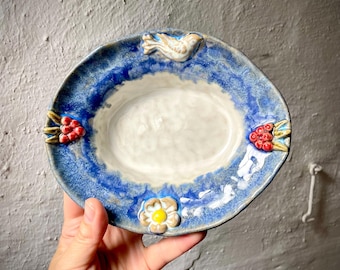 Puede incluir: Un plato ovalado de cerámica con centro color crema y borde azul texturizado. El borde está decorado con un pequeño pájaro esculpido en la parte superior, una flor blanca con centro amarillo en la base y racimos de bayas rojas a los lados.