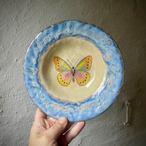 Könnte beinhalten: Eine runde Keramikschale mit cremefarbenem Zentrum, das einen gemalten Schmetterling mit gelben, rosa und blauen Flügeln zeigt. Die Schale hat einen breiten, strukturierten Rand in Blau- und Weißtönen mit gesprenkelter Optik und unebenen Kanten.