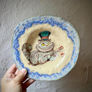 Könnte beinhalten: Eine runde Keramikschale mit cremefarbener Mitte, die eine handgemalte Illustration einer grinsenden Katze mit einem blaugrünen Zylinder mit rotem Band zeigt, umgeben von kleinen roten Pilzen. Die Schale hat einen strukturierten, blau-weißen Rand.
