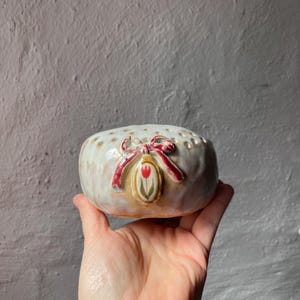 Könnte beinhalten: Eine kleine, cremefarbene Keramikschale mit strukturierter Oberfläche und kleinen Löchern am oberen Rand. Die Schale hat eine rote Schleife und ein Tulpenmuster. Die Schale wird in einer Hand gehalten.