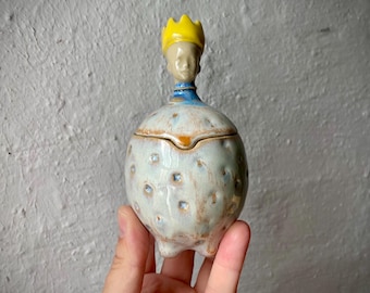 Puede incluir: Un tarro de cerámica con tapa, con un esmalte texturizado de color blanco roto y pequeñas hendiduras. La tapa está rematada con una figura esculpida que lleva una corona amarilla brillante y un cuello azul, con un rostro de tonos neutros.