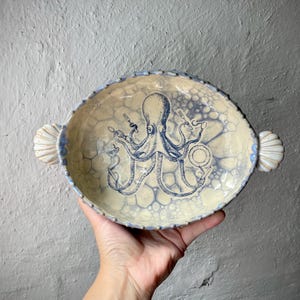 Könnte beinhalten: Eine ovale Keramik-Servierschale mit einer cremefarbenen und blauen Glasur im Blasenmuster. In der Mitte befindet sich eine detaillierte blaue Tintenfisch-Illustration, die Besteck hält. Die Schale hat zwei muschelförmige Griffe und einen Perlenrand.