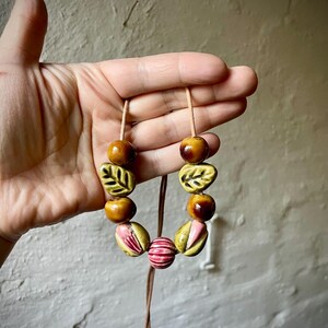 Puede incluir: Un collar artesanal con cuentas de cer&aacute;mica en tonos marr&oacute;n, rosa y verde. El collar incluye cuentas en forma de hoja y redondas, ensartadas en un cord&oacute;n marr&oacute;n. El dise&ntilde;o es terroso y org&aacute;nico.