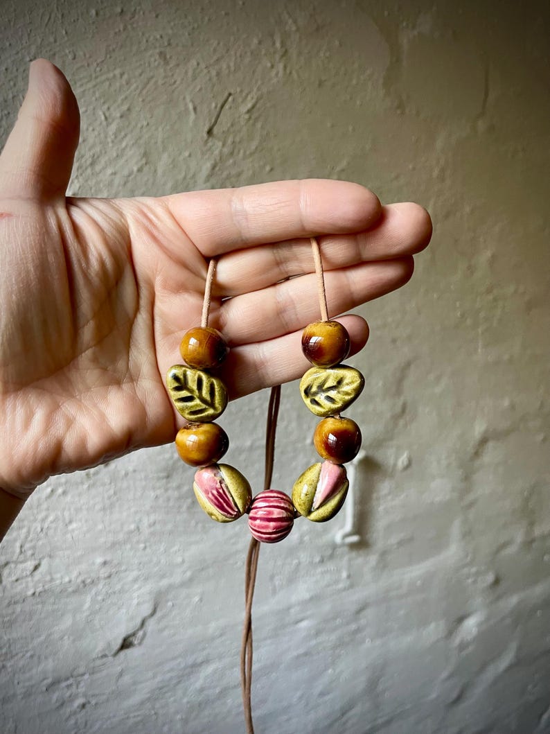 Puede incluir: Un collar artesanal con cuentas de cer&aacute;mica en tonos marr&oacute;n, rosa y verde. El collar incluye cuentas en forma de hoja y redondas, ensartadas en un cord&oacute;n marr&oacute;n. El dise&ntilde;o es terroso y org&aacute;nico.
