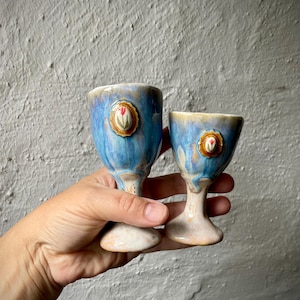 Puede incluir: Dos cálices de cerámica con un esmalte brillante en azul y crema. Cada cáliz presenta un pequeño medallón decorativo de forma ovalada en la parte frontal, que muestra un tulipán rojo pintado sobre fondo blanco con un borde dorado festoneado.