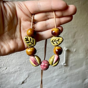 Puede incluir: Un collar hecho a mano con cuentas de cer&aacute;mica en tonos marr&oacute;n, verde y rosa. Las cuentas incluyen formas redondas, dise&ntilde;os de hojas y patrones a rayas, ensartadas en un cord&oacute;n de cuero. Un accesorio art&iacute;stico &uacute;nico.