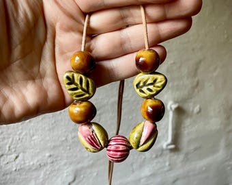 Puede incluir: Un collar hecho a mano con cuentas de cerámica en tonos marrón, verde y rosa. Las cuentas incluyen formas redondas, diseños de hojas y patrones a rayas, ensartadas en un cordón de cuero. Un accesorio artístico único.