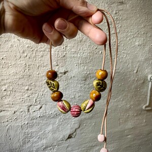 Puede incluir: Un collar hecho a mano con cuentas de cer&aacute;mica en tonos marr&oacute;n, verde y rosa. Las cuentas est&aacute;n ensartadas en un cord&oacute;n beige, con dos cuentas de cer&aacute;mica rosas en los extremos. El collar se muestra contra una pared blanca texturizada.