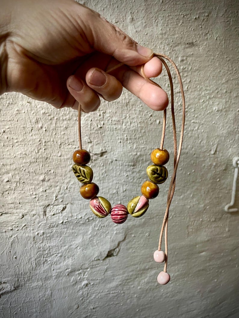Puede incluir: Un collar hecho a mano con cuentas de cer&aacute;mica en tonos marr&oacute;n, verde y rosa. Las cuentas est&aacute;n ensartadas en un cord&oacute;n beige, con dos cuentas de cer&aacute;mica rosas en los extremos. El collar se muestra contra una pared blanca texturizada.