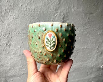 Puede incluir: Una taza de cerámica verde con una superficie texturizada y abultada, con un medallón ovalado central que presenta una ilustración pintada de una flor roja con hojas azules sobre un fondo blanco. El esmalte tiene motas marrones en el acabado.