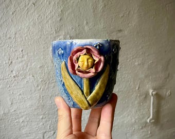 Puede incluir: Una taza de cerámica con esmalte azul texturizado que presenta una flor rosa esculpida en tres dimensiones con una cara amarilla en el centro y dos hojas verdes que se extienden hacia arriba desde la base de la flor sobre el fondo azul.