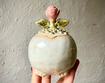 Puede incluir: Un tarro de cerámica redondo con un esmalte brillante, pálido e iridiscente. La tapa presenta un capullo de flor rosa esculpido con dos hojas verdes sobre una superficie texturizada y perlada. La base del tarro tiene pequeños pies redondeados y el acabado general muestra variaciones sutiles en color y textura.