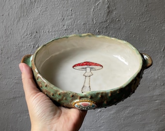 Puede incluir: Un cuenco de cerámica redondo con dos pequeñas asas, que presenta un exterior verde texturizado y un interior liso de color crema. En el centro hay una ilustración detallada de una seta de sombrero rojo y un pequeño motivo floral en el borde.