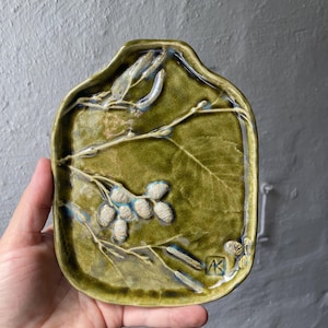 Könnte beinhalten: Handgefertigte, olivgrüne Keramikschale mit rechteckiger Form und abgerundeten Kanten. Die Schale zeigt ein erhabenes botanisches Design aus Blättern und Samenkapseln in Weiß und Blau. Die Initialen des Künstlers sind sichtbar.