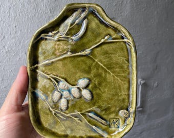 Puede incluir: Un plato de cerámica verde oliva hecho a mano, de forma rectangular con bordes redondeados. El plato presenta un diseño botánico en relieve de hojas y vainas de semillas en blanco y azul. Las iniciales del artista son visibles.