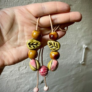 Puede incluir: Un collar hecho a mano con cuentas de cer&aacute;mica en tonos marr&oacute;n, verde y rosa. Las cuentas incluyen formas de hojas, cuentas redondas y cuentas rayadas, ensartadas en un cord&oacute;n marr&oacute;n claro. El collar est&aacute; sostenido por una mano.