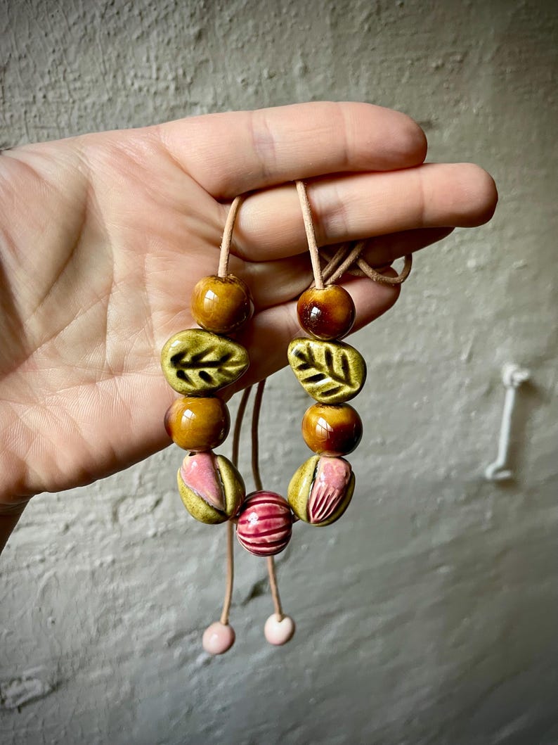 Puede incluir: Un collar hecho a mano con cuentas de cer&aacute;mica en tonos marr&oacute;n, verde y rosa. Las cuentas incluyen formas de hojas, cuentas redondas y cuentas rayadas, ensartadas en un cord&oacute;n marr&oacute;n claro. El collar est&aacute; sostenido por una mano.