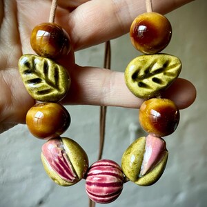 Puede incluir: Collar hecho a mano con cuentas de cer&aacute;mica de varias formas y colores. El collar incluye esferas marrones brillantes, cuentas en forma de hoja verde oliva y cuentas a rayas rosas y rojas, ensartadas en un cord&oacute;n beige.
