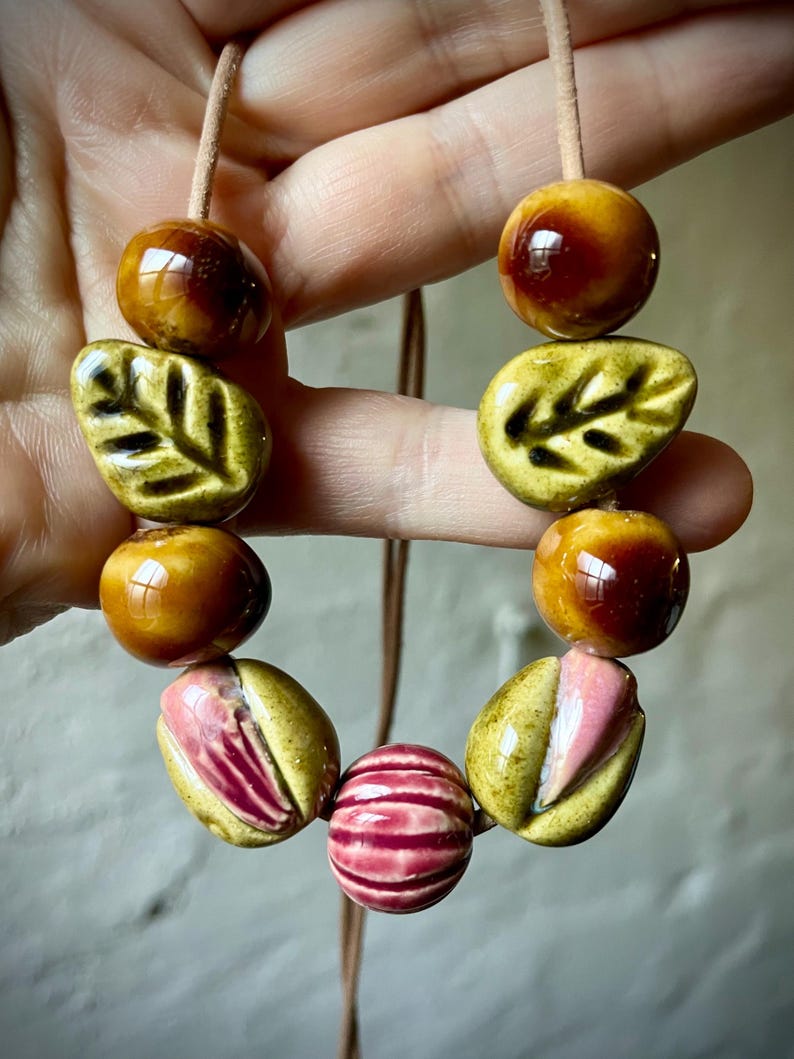 Puede incluir: Collar hecho a mano con cuentas de cer&aacute;mica de varias formas y colores. El collar incluye esferas marrones brillantes, cuentas en forma de hoja verde oliva y cuentas a rayas rosas y rojas, ensartadas en un cord&oacute;n beige.