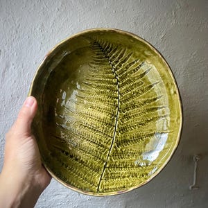 Könnte beinhalten: Eine runde Keramikschale mit einer glänzenden olivgrünen Glasur. Das Innere zeigt ein detailliertes, geprägtes Farnblattmuster, das sich über den gesamten Boden erstreckt, mit einem sichtbaren Riss, der vertikal durch die Mitte des Blattabdrucks verläuft.
