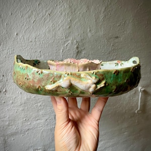 Op de afbeelding: Een ondiepe keramische kom met een groen gevlekte buitenkant met een reliëf van een rennend konijn en kleine pootafdrukken. De binnenkant heeft een bleek oppervlak met bloemaccenten en een centraal roze element met geschulpte randen.