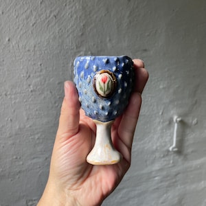 Könnte beinhalten: Ein blauer Keramik-Eierbecher mit einer texturierten, noppigen Oberfläche und einem weißen Standfuß. Auf der Vorderseite befindet sich eine kleine, ovale, dekorative Einlage, die eine rote Tulpe mit grünen Blättern auf weißem Grund zeigt.