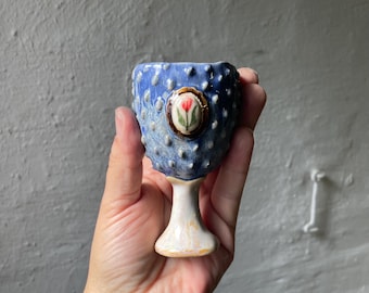 Puede incluir: Un huevero de cerámica azul con una superficie texturizada y abultada y una base de pedestal blanca. La parte frontal presenta una pequeña incrustación decorativa ovalada que representa un tulipán rojo con hojas verdes sobre un fondo blanco.