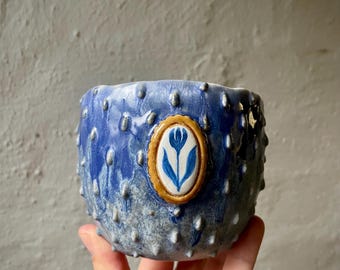 Puede incluir: Una taza de cerámica con una superficie texturizada y abultada que presenta un esmalte degradado en azul y blanco. El centro de la taza muestra un medallón ovalado con un diseño de tulipán azul pintado a mano en relieve y borde dorado.